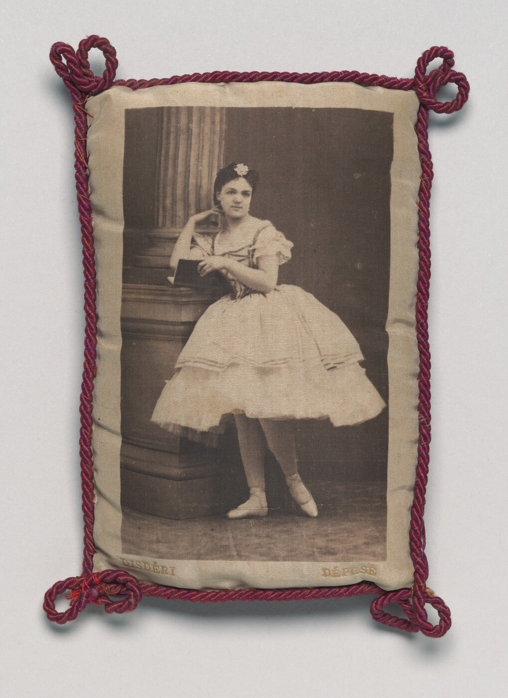 The image displays a woman in a ballet outfit, standing en pointe next to a column. She has delicate facial features, neatly styled hair with a floral headpiece, and a serene expression. The woman wears a classic ballet costume with a fitted bodice, short puffed sleeves, and a full tulle skirt. She is holding a book and is surrounded by a stage or studio setting with a classical column. The image is framed with twisted red cords.