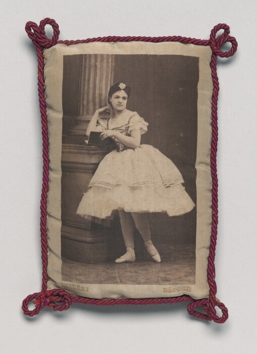 The image displays a woman in a ballet outfit, standing en pointe next to a column. She has delicate facial features, neatly styled hair with a floral headpiece, and a serene expression. The woman wears a classic ballet costume with a fitted bodice, short puffed sleeves, and a full tulle skirt. She is holding a book and is surrounded by a stage or studio setting with a classical column. The image is framed with twisted red cords.