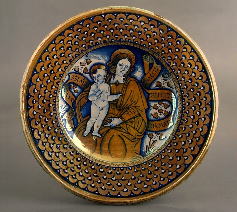 The image depicts a circular ceramic plate featuring a painted illustration at its center. The artwork shows the upper body and torso of an adult figure seated in a frontal position, holding a young child. The adult has large eyes and hair styled in two bunches above their ears. Their hair is reddish-brown, with a circular halo above their head. The adult wears a brown robe with a blue outline. The child, shown completely naked and standing, has light-colored hair and a halo, with one hand raised. The surrounding design includes floral motifs and scrolling banners inscribed with text. The plate's outer rim has a pattern of overlapping blue and golden scales.