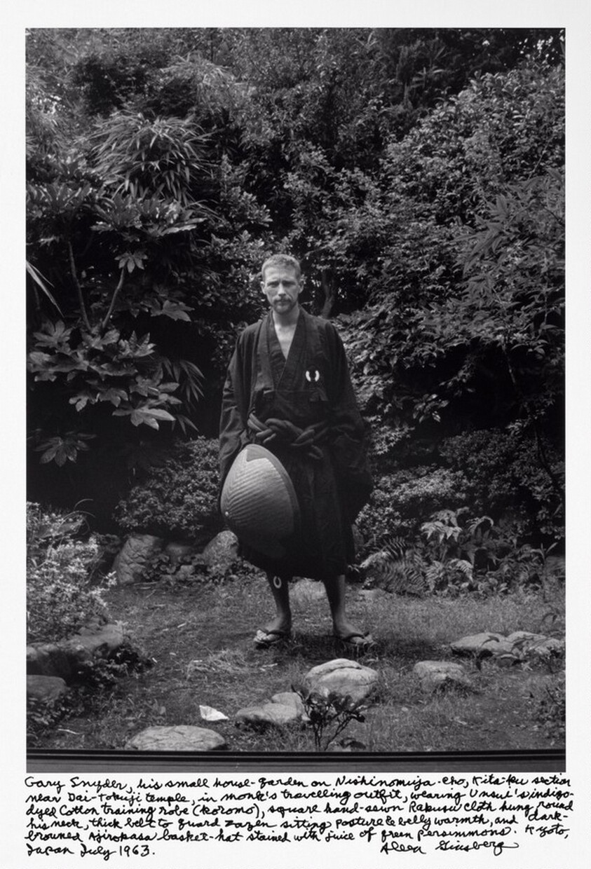 Gary Snyder, his small house-garden on Nishinomiya-cho, Kita-ku section near Dai-tokuji temple, in monk’s travelling outfit, wearing Unsui’s indigo-dyed cotton training robe (koromo), square handsewn Rakusu cloth hung round his neck, thick belt to guard zazen sitting posture & belly warmth, and dark-browned Ajirokasa basket-hat stained with juice of green persimmons. Kyoto, Japan July 1963.