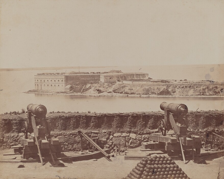 The image shows a view of gun emplacements with cannons pointed towards a fort across a body of water. The horizon is low, almost blending with the fortress in the distance. The fort is positioned off-center to the right, showcasing its stone structure by the water. Two cannons are mounted on wooden platforms in the foreground, alongside a stack of cannonballs. The color palette is monochrome with sepia tones, giving it an aged look. The landscape is barren with minimal vegetation, suggesting a utilitarian military environment.