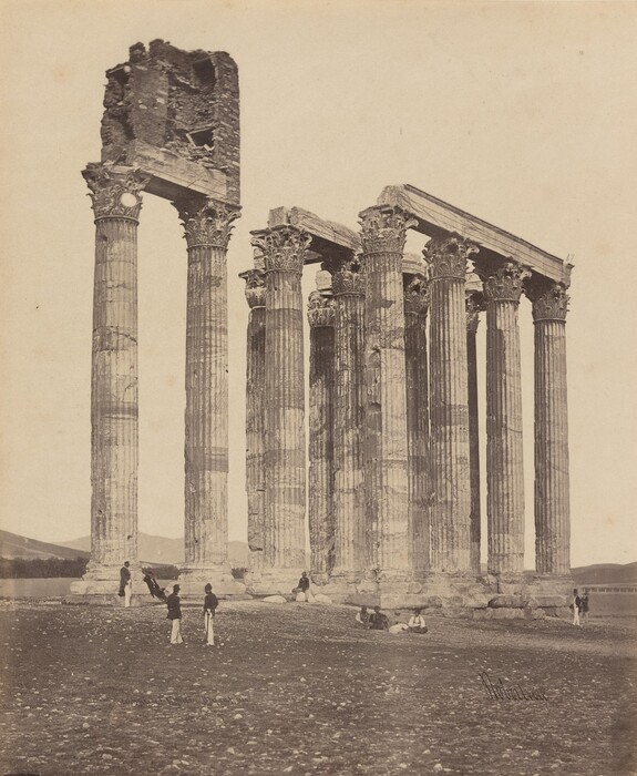 The image shows a view of ancient Doric columns, part of a classical ruin. There are eight visible columns with intricate capitals and detailed carvings. The columns support a damaged block. In the background, there are distant hills. A group of men is dispersed near the columns on a barren, rocky ground. Some are standing while others are seated. Subdued bushes are also visible in the image, hinting at the arid environment surrounding the ruins.