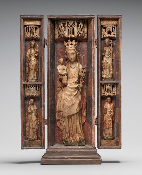 This freestanding sculpture consists of a central panel with two narrower panels on hinges that could close like shutters. The central panel shows a crowned woman holding a baby, Mary and Jesus, and each side panel holds two people, one stacked over the other and about half the scale of Mary. All five people are shown under canopies carved with triangular moldings carved with filigree-like tracery. The panels appear to be wood, and the people are carved from white alabaster. The faces are all oval with large eyes, straight noses, and small mouths. They all wear robes, dresses, or habits that fall in thick folds to the small bases on which they stand. Mary has long, wavy hair, and she holds a sprig of roses in her left hand, to our right. Resembling a miniature man, Jesus sits upright in her other hand, his hands clasped in his lap and his knees together. He appears to wear a collared vestment, and his hair is either damaged or is curly only on each side of his narrow face. To the left, the top woman wears a crown over long wavy hair. She props the end of a chest-high sword with one hand and holds a small wheel with the other. Below her, a woman wears a cloth covering over her head and a rosary hangs from her waist. She holds a what might be small vessel and a green object, perhaps a book. In the top right, the woman also has wavy hair under a crown, and she holds a book and a tall cross. The fifth woman, in the lower right, holds two objects that are difficult to make out, perhaps an incense burner or jar and a short staff. Traces of red, green, black, and other colored paint are especially prominent on the objects the women hold and the backgrounds.