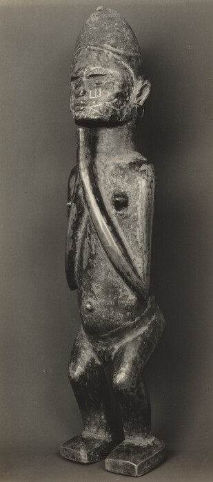 The sculpture portrays a humanoid figure standing upright with hands together under the chin. The figure is depicted as a male with minimal anatomical details, wearing a conical hat. The face has closed eyes, an exaggerated nose, and vertical marks on the cheeks. It is made of wood with a weathered appearance, showing a visible grain. The figure has hoop earrings and large, flat feet for stability, with elongated proportions.