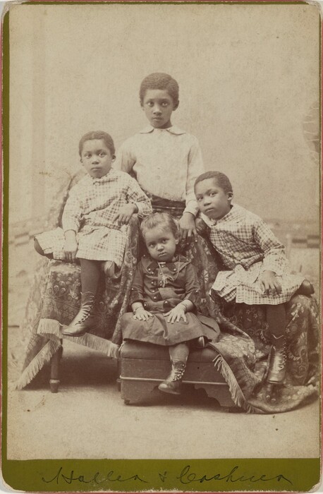 The image shows four young children. The central child standing at the back wears a light-colored shirt. Flanking the central child are two children sitting on a cushioned seat draped with fringed fabric. Both have short, textured hair and wear patterned dresses, stockings, and laced boots. The child on the right rests their arm on the seat, with legs crossed and hands on their knees. The child seated in the front wears a darker dress with lace detailing and has curled hair styled into a top bun with ribbons or a bow. This child wears bracelets and looks slightly away from the camera. The children are surrounded by ornately patterned fabric covering the furniture and a textured backdrop, likely in a photography studio.