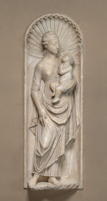 Carved in high relief in white marble, a woman and the baby she holds are framed within a curved arch. The woman’s body faces us, but she turns her face to look down at the baby tucked into her left arm, to our right. The woman’s long hair is pulled back under a twisted band, and a few tendrils fall on her face. Her nose is pointed, and she has full lips over a narrow chin. Her dress is fastened on the chest with a button or brooch and is tied across the chest and high around the waist. The garment has elbow-length sleeves, and it falls in a swag across her hips over a long skirt that stops just short of her sandaled feet. The fabric is sheer so the nipple we can see is visible, and the fabric gathers in fine, shallow folds. Her other elbow is bent and held by her side, and the fingers on both hands are noticeably long. The child turns a chubby face up to the woman and crosses his hands over his chest. He is covered only by a cloth across his middle, and he has short, wavy hair. Pudgy rolls make creases on his neck, arms, and especially chunky thighs. The niche in which they stand has a fluted shell in the rounded portion above molding. The remaining niche is smooth, and the entire sculpture is set into a tan-colored wall.