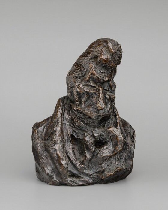 The sculpture depicts a human bust, capturing the head and shoulders of the subject. The figure is artistically represented with rough, textured features including pronounced facial elements and an exaggerated coiffure that rises prominently on the head. The sculpture is predominantly a dark bronze color with subtle variations in shade that highlight its rugged surface. Made of bronze, the texture of the sculpture's material gives it a weathered appearance, with visible ridges and grooves adding depth. The subject appears clad in a coat with a broad lapel and tied neckwear, articulated through exaggerated, abstract forms that merge into the rest of the bust. The overall appearance is reminiscent of classic portrait busts, elevated in a modern, abstract style, giving it a solid and distinguished presence relative to a viewer.