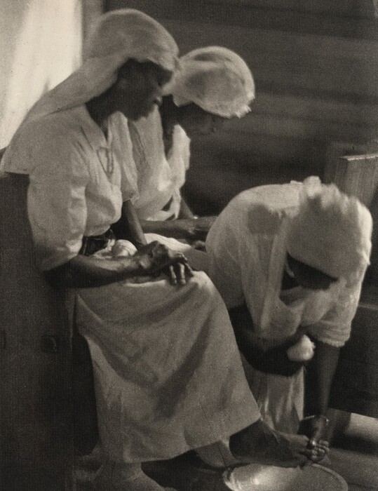 The image shows three women participating in a foot-washing ritual. The women are partially depicted, focusing on their upper bodies and the action of two women washing another woman's foot. They are sitting close to one another with two of them leaning forward to wash. Their faces are partly obscured by shadows. Each woman wears a light-colored headscarf and a white dress. The woman whose foot is being washed sits upright while the others lean over a basin on the floor. The setting is indoors with a wooden background visible, creating an intimate, softly lit atmosphere.