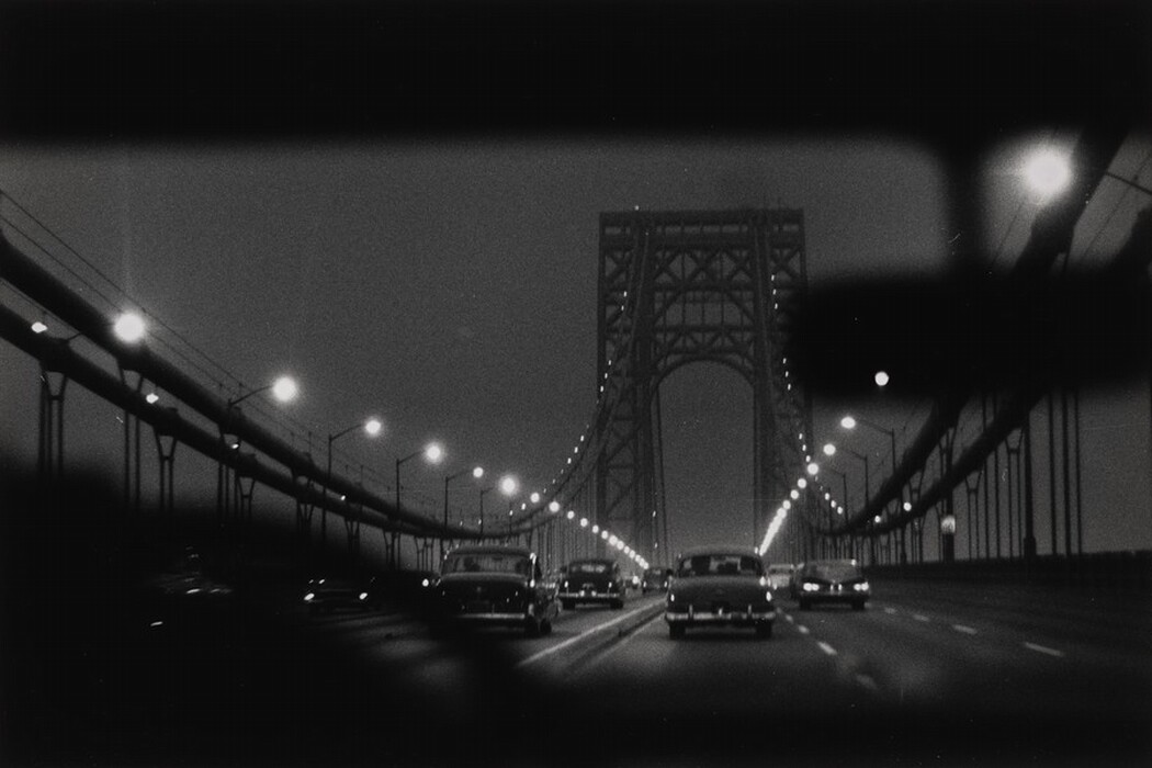 The image shows a view from inside a car traveling on a bridge with several cars heading in the same direction. The bridge is suspended between two metal structures, with streetlights along its length creating a line of glowing orbs. The steel framework of the bridge stretches into the distance, and the horizon is obscured by its architectural elements. The scene is captured in black and white, emphasizing the bridge's silhouette against a faintly illuminated sky. Smooth, fluid lines depict the bridge and vehicles, creating a strong graphic contrast in shades of grey and black.