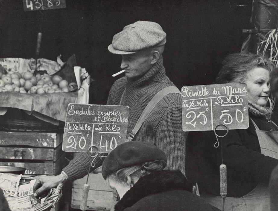 This is a photograph of a market scene. The image shows a bustling marketplace with various produce items visible in the background. At the center of the image, there is a display with hand-written signs showing prices for different products. The atmosphere is lively, with goods arranged in crates and baskets, some covered with paper. The image emphasizes the traditional and vibrant nature of local market commerce.
