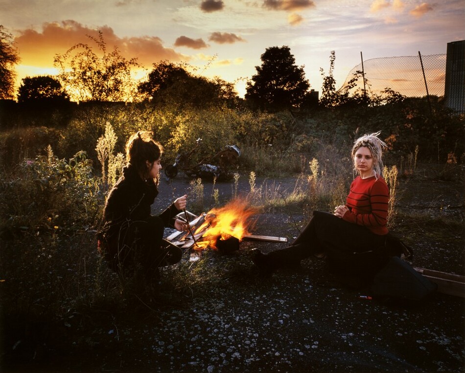 The image depicts two people outdoors, seated on the ground. One person has dark hair tied up, crouching near a small fire. The other person, with light blonde dreadlocks, faces the camera. The first person wears dark clothing while the second person wears a red shirt with black stripes. The background includes greenery, a wire fence, and an orange-hued sunset sky.