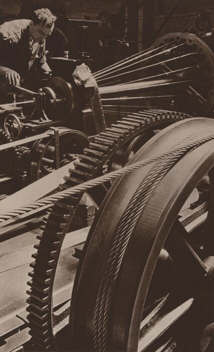 This is a sepia-toned photograph showing machinery. There are large gears and ropes or cables, with a sizable gear wheel on the right side. The ropes extend outward diagonally and converge at a central point. A person appears to be working on the machine in the upper left, leaning forward and touching a component with his hands. The background contains additional mechanical elements and shadowed areas.