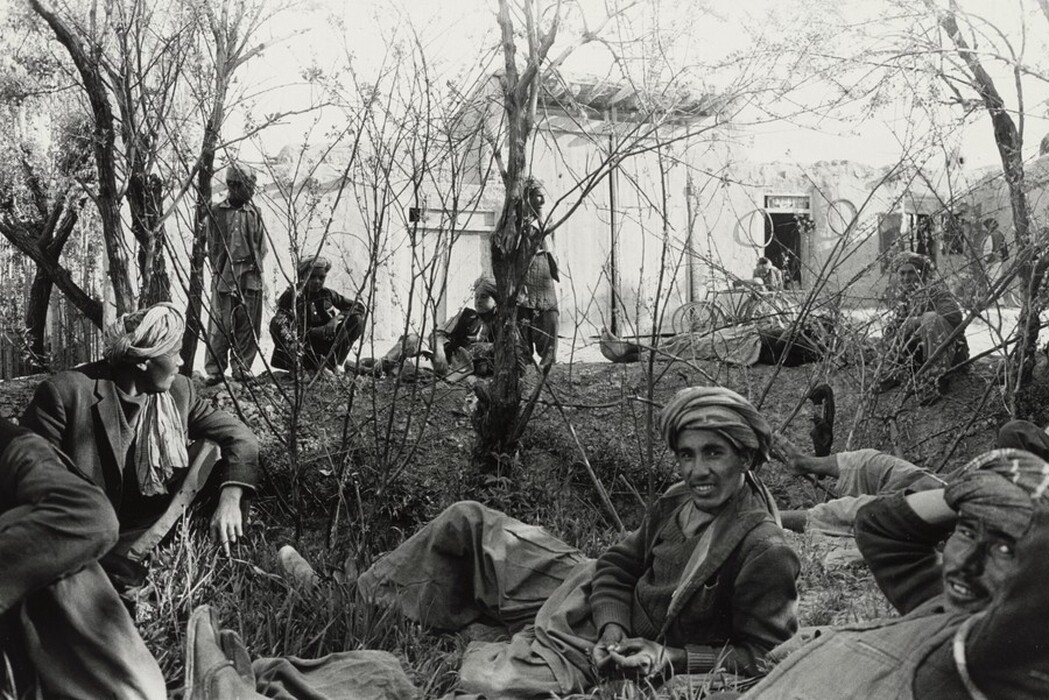 The image shows several men outdoors in a natural setting. One man is lying on the ground facing the camera, supported by his elbow, wearing a headscarf, sweater, and loose trousers. Another man in a suit jacket and headscarf is sitting nearby, leaning back with his arm extended for support. Other men are dressed in loose garments and are either sitting or lying on the ground under sparse branches of small trees or shrubs. In the background, there is a simple building with a flat roof, window openings, and possibly a door, with a bicycle leaning against it.