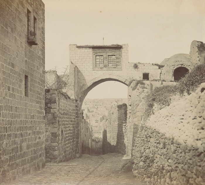 The image shows a view of a narrow, cobbled lane between stone walls. An archway with a small structure on top is slightly off-center to the right. Stone buildings can be seen in the distance. The color palette is muted and natural, typical of aged stone. The texture of the stonework is detailed, adding to the historic feel.