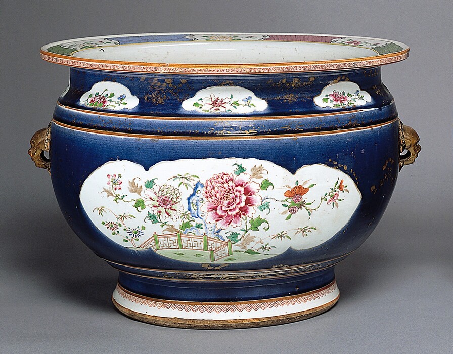 This is a photograph of a ceramic vessel. The vessel is primarily dark blue with white panels that are decorated with floral patterns and scenes, featuring pink, red, and green flowers. The upper rim and base of the piece are also white with floral designs and gold accents. The vessel is equipped with decorative golden handles.