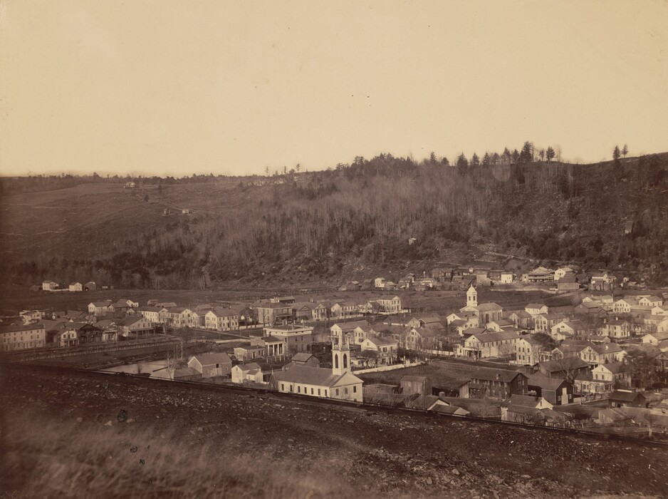 The image shows a small town in a valley, viewed from an elevated perspective. The horizon is marked by hills with trees, and the town has various buildings with different rooflines. A railway track is visible in the foreground. The color scheme is warm with sepia tones, giving the image a vintage look.