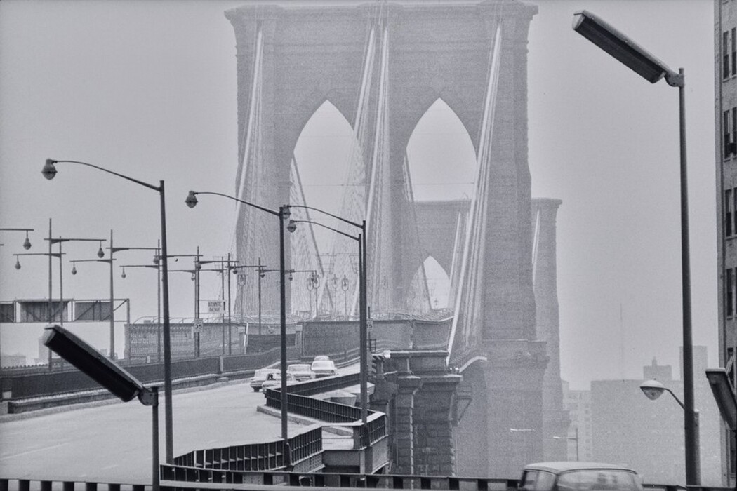 The image shows the Brooklyn Bridge from a slightly elevated position. The horizon, bridge structure, cityscape, cars on the road, street lamps, signs, and the black-and-white color scheme are visible.