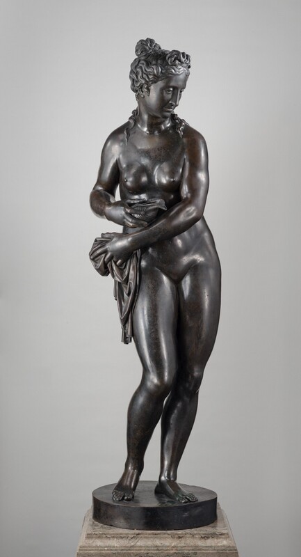 This free-standing bronze sculpture is of a nude woman who reaches her left arm, on our right, across her body to hold a piece of drapery bunched at her opposite hip, while she holds a conch seashell in her other hand. In this photograph, her body faces us and she looks down and to our right. She has a straight nose, and her full lips are closed. Her long, wavy hair is gathered into a knot at the top of her head, and a few tendrils trail down her neck and shoulders. Her sloping shoulders frame round breasts, which are placed close together on her chest. Most of her weight is on her left leg, on our right, and her other knee is bent so she balances on the ball of that foot. This means her hips sway slightly to our right as her arms sweep to our left. She stands on a round, disk-like plinth on a veined, tan marble base. Light glints off the polished, deep brown bronze surface of the sculpture.