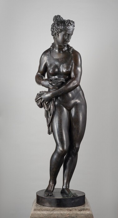 This free-standing bronze sculpture is of a nude woman who reaches her left arm, on our right, across her body to hold a piece of drapery bunched at her opposite hip, while she holds a conch seashell in her other hand. In this photograph, her body faces us and she looks down and to our right. She has a straight nose, and her full lips are closed. Her long, wavy hair is gathered into a knot at the top of her head, and a few tendrils trail down her neck and shoulders. Her sloping shoulders frame round breasts, which are placed close together on her chest. Most of her weight is on her left leg, on our right, and her other knee is bent so she balances on the ball of that foot. This means her hips sway slightly to our right as her arms sweep to our left. She stands on a round, disk-like plinth on a veined, tan marble base. Light glints off the polished, deep brown bronze surface of the sculpture.