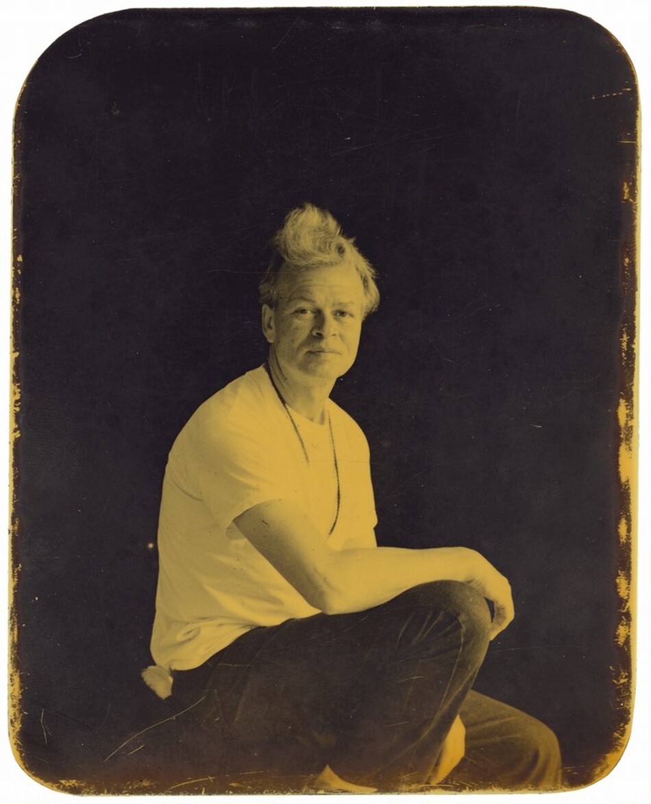The image shows a person from the waist up, in a three-quarter view. The individual is seated, with one arm resting on their knee and facing slightly to the left. They have light, textured hair styled upwards. Their facial expression is calm, with light shading on their face. The person is wearing a light-colored t-shirt, dark pants, and a thin necklace. The background is dark, highlighting the subject. The image has rounded corners and a sepia tone.