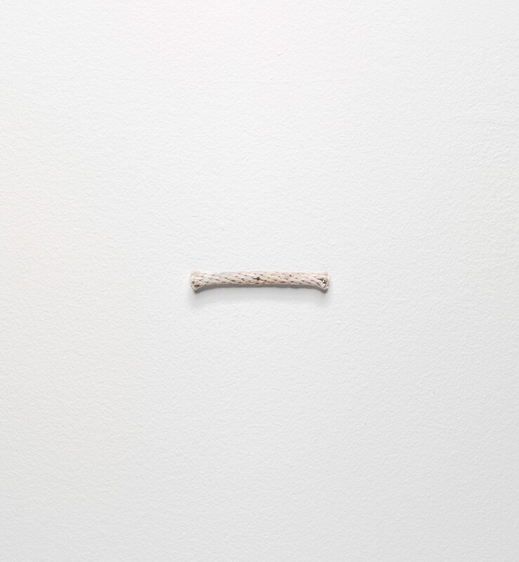 A short length of ivory-white, braided rope is nailed horizontally on a white wall in this square photograph. Pin-like nail heads are visible at the center, and at the left and right edges of the rope.