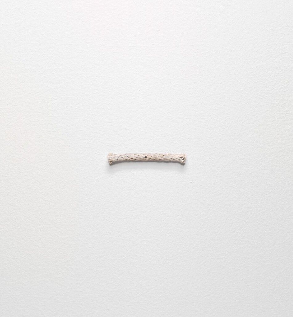 A short length of ivory-white, braided rope is nailed horizontally on a white wall in this square photograph. Pin-like nail heads are visible at the center, and at the left and right edges of the rope.