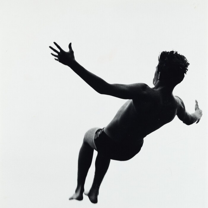 The image shows a man jumping in mid-air with his entire body visible from behind. He appears to be leaning to one side with both arms extended, one arm higher than the other. The man's facial features are not visible due to the angle, but his hair is short and dark. He is wearing dark swim trunks, and there are no objects or jewelry visible on him. The background is a stark white.