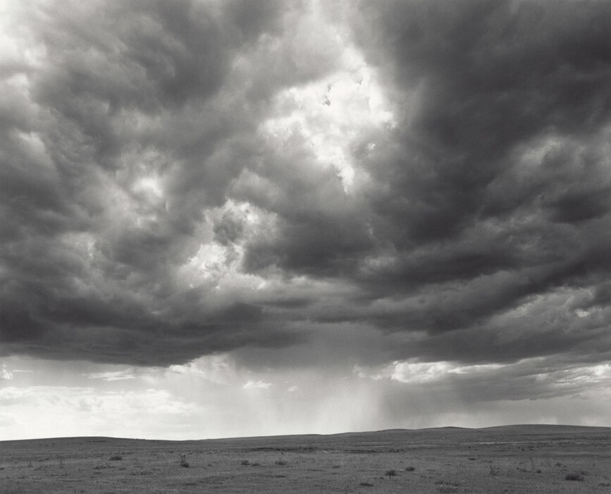 The image shows a wide landscape under a cloudy sky. The horizon is low in the frame, separating the land from the sky with subtle undulations. Dark clouds fill the sky, with varying shades of gray. Some clouds are very dark, suggesting rain, while lighter patches indicate breaks in the overcast. The color palette is predominantly gray. The land is sparsely vegetated, accentuating the starkness of the landscape.