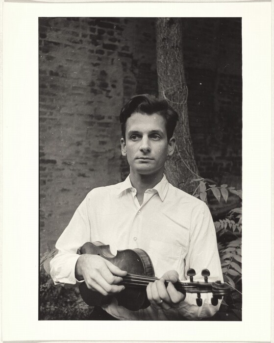 The image depicts the upper half of a man standing upright. His face is visible, showing a clean-shaven face, straight nose, and focused expression. His hair is dark with a slight wave, styled back from the forehead. He wears a light-colored shirt with a collar, buttoned but without a tie. He holds a violin, with his left hand on the neck and his right hand on the body. The background shows a brick wall, a tree trunk, and foliage, suggesting an outdoor setting.