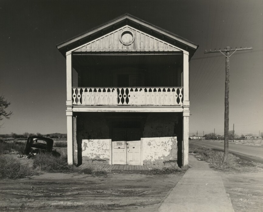 The image is a frontal view of a two-story wooden building with a gabled roof, decorative elements, and a balcony with a geometric railing design. There are two boarded-up doors at the lower level. Utility poles line a road that extends into the distance on the right side, with an old partially visible vehicle on the left. The sky is clear in this black and white photograph, creating stark contrasts. Shadows are sharp, depicting the texture and aging of the structure and surroundings.