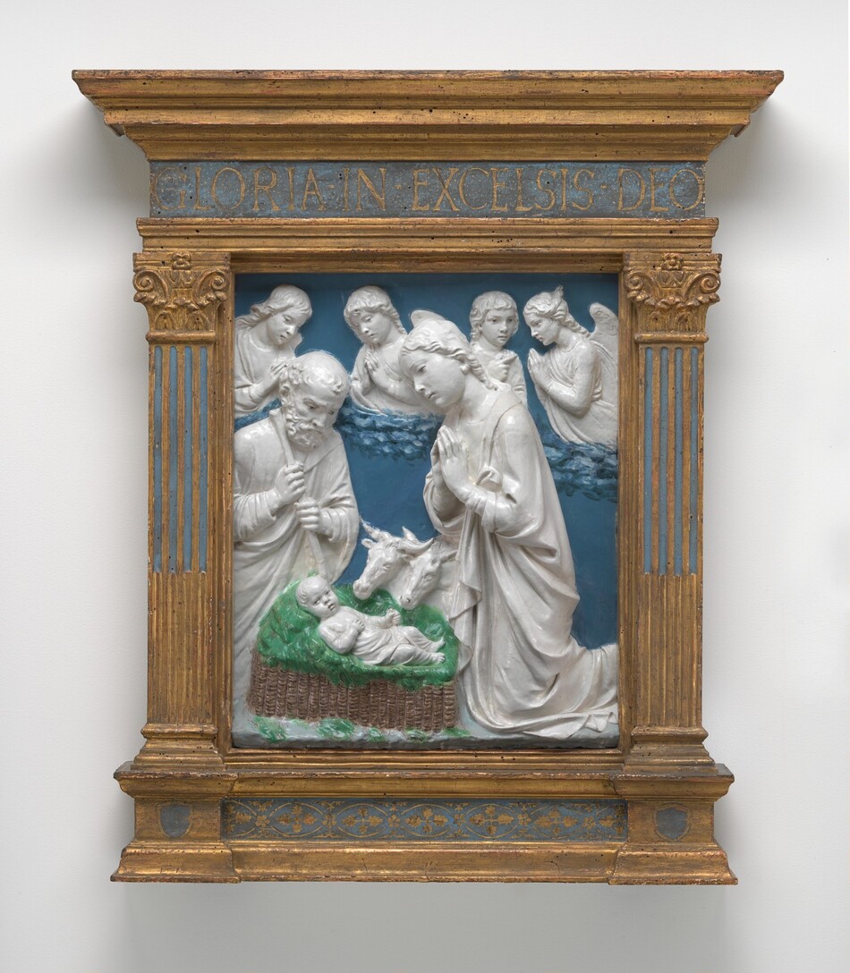 The sculpture is a relief depicting a scene with people gathered around a baby in a basket, surrounded by green grass. Surrounding the basket, there is a man on the left and a woman on the right with her hands clasped in prayer. Behind them is a row of more people, including two angels. Two oxen or cattle can be seen looking down at the child. The people and animals are all white against a deep blue backdrop. The sculpture is framed within an intricate gold-and-blue frame, with the words "GLORIA IN EXCELSIS DEO" inscribed at the top. The golden frame has detailed columns with floral and geometric patterns.