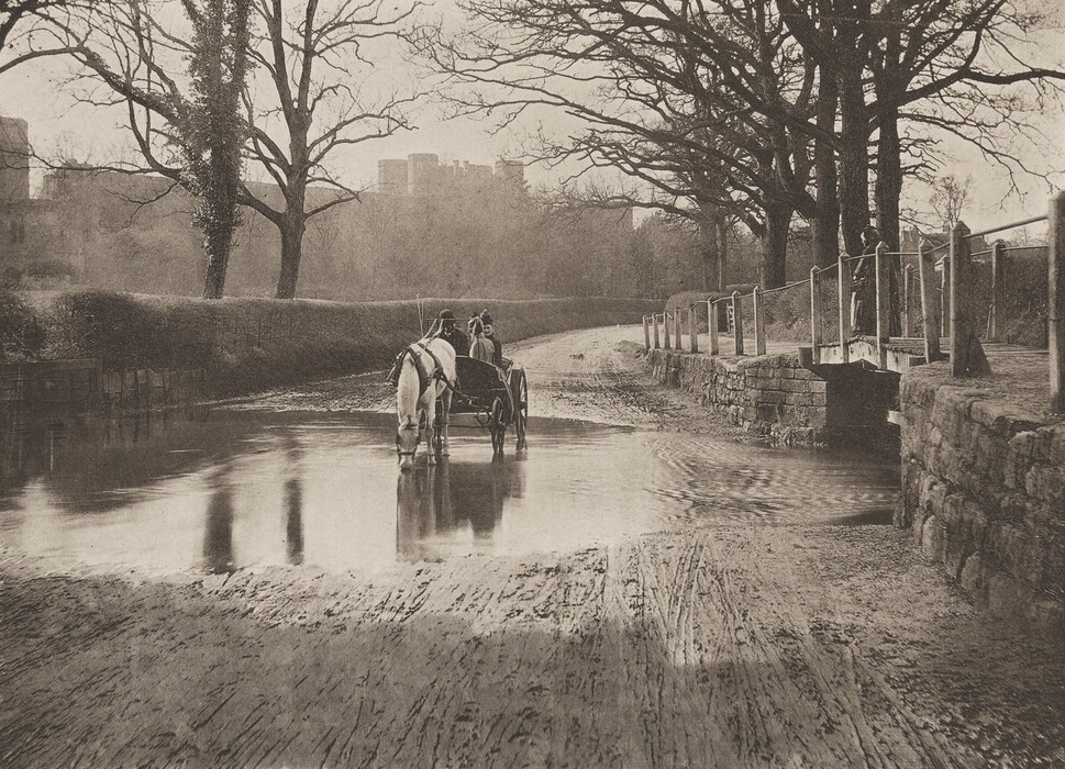 The image shows a low vantage point of a tranquil scene by a shallow water crossing. An earthen path with wheel and foot marks leads to the water, where a horse-drawn carriage is stopping. A horse drinks from the water, its reflection visible. In the background, a dirt road curves to the right, bordered by a stone wall and wooden fence. Leafless trees with intricate branches line the road, leading to a large structure, possibly a castle, faintly visible in the mist. The horizon is marked by distant trees blending into the gray sky. The color palette is earthy and subdued.