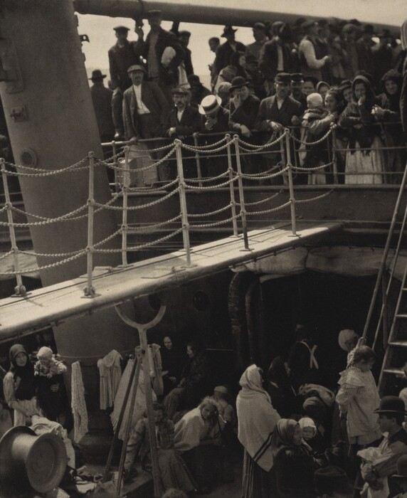 This is a photograph of passengers aboard a ship. It captures a large group of individuals gathered on the decks of the ship. The scene includes rope barriers, ladders, and railings. The lighting and sepia tone suggest a snapshot from a past era. The deck is filled with activity; some individuals sit or lean against the ship’s structure, while others stand or look out from the upper level. The composition conveys a sense of journey and migration.