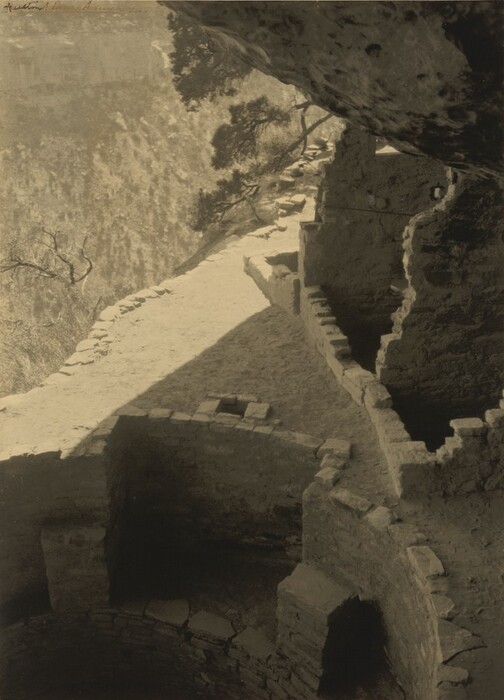 We look sharply down at the ruins of stone walls and structures tucked under an overhanging rock close to us, as the land drops vertiginously away in this vertical black and white photograph. Some of the walls are straight and some curved to follow the contours of what appears to be a cliff face. A few scrubby branches grow from the rock at the top center of this composition. We look almost straight down onto a tree-filled canyon to our left.