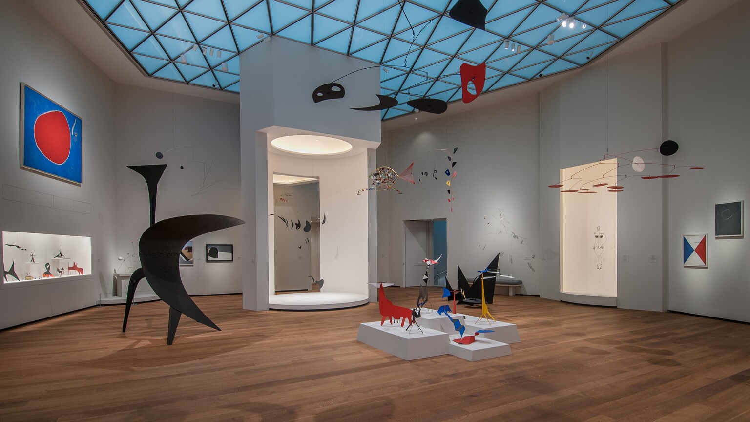 A room with white walls, hardwood floors, and skylights, with various sculptural forms, mobiles, and some paintings on display.