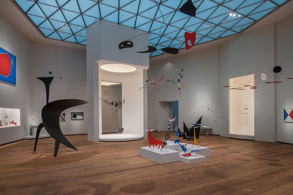 A room with white walls, hardwood floors, and skylights, with various sculptural forms, mobiles, and some paintings on display.