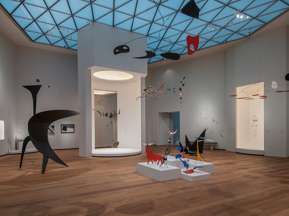A room with white walls, hardwood floors, and skylights, with various sculptural forms, mobiles, and some paintings on display.