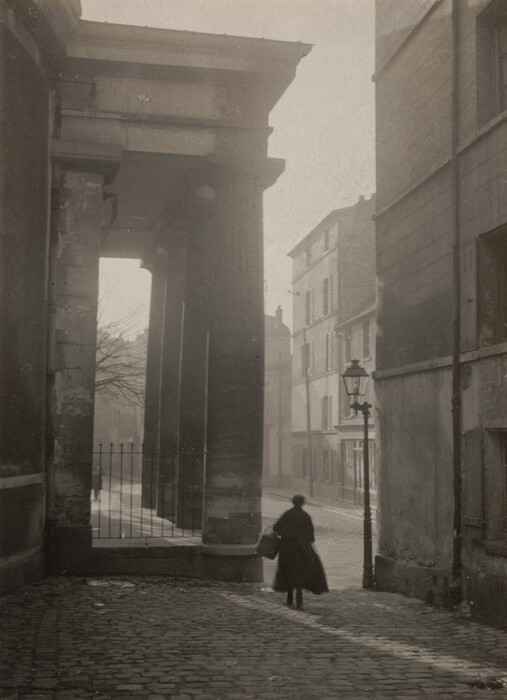 This is a photograph of an empty cobblestone street with large columns and buildings. The image captures an urban scene where a street winds between tall columns on the left and a building on the right. Sunlight casts long shadows on the ground, adding depth to the image. In the background, more buildings line the street, while a lamppost stands on the sidewalk. The atmosphere is serene, with light fog diffusing the sunlight.