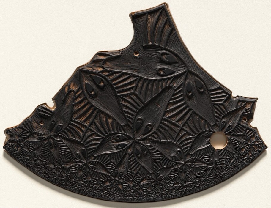 The sculpture is fan-shaped and adorned with intricate carvings depicting stylized leaf-like forms with eye-shaped cutouts. It is made of a dark material, likely wood, with a blackened finish revealing a lighter color underneath. The carvings present overlapping leaf-like forms in a dense pattern, creating a textured appearance. The object appears flat with detailed etchings that play with light and shadow. Small holes along the edges may serve as attachment points or decorative accents.
