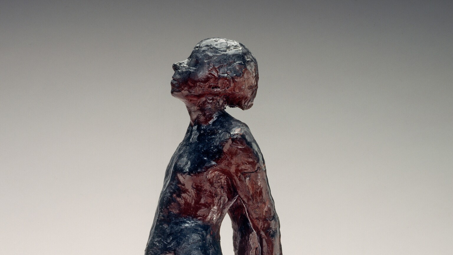 A young, nude girl is roughly modeled with glossy navy blue and wine-red beeswax in this freestanding sculpture. She faces our left in profile in this photograph. Her weight rests on her left foot, closer to us, and her other leg extends long, the foot turned out at an angle. Her hands are clasped behind her back, and her round belly projects from her swayed hips. Her head is tipped back and her eyes closed. She stands on a double pedestal. A crack runs across one corner of the top, white, square plaster pedestal. A wider wood pedestal is below.