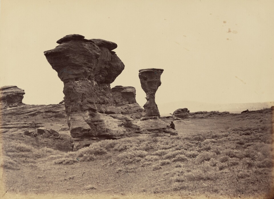 The image depicts a rugged landscape with eroded rock formations dominating the scene. The viewpoint is at ground level, looking up at the towering rocks. The horizon extends into the distance with a hazy sky. The color palette is monochromatic brown. Sparse shrubbery covers the terrain, contrasting with the rock formations. A small figure sits on the rocks near the center, providing scale.