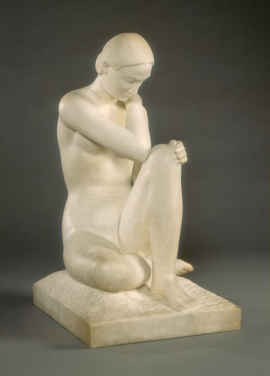 The sculpture is of a seated nude figure in a contemplative pose. The individual is sitting with one knee bent and the arms folded over it, seeming to rest their head gently against the knee. The figure is carved from light-colored marble, with a smooth and polished surface. The marble gives the sculpture a slight sheen, highlighting the details and curves of the figure's form. The sculpture portrays a realistic representation of the human figure, capturing natural proportions and anatomical details.