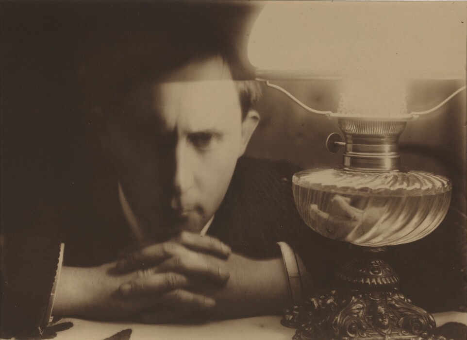 The image shows a man's upper body, focusing on his face and hands as he leans forward on a table. His face is partially shaded, showing a serious expression with deep-set eyes and a slightly furrowed brow. He has short, dark hair and is dressed in a dark suit with a white shirt visible at the cuffs and collar. An ornate lit oil lamp with a glass body and metal base is next to him on the table. The background is dark, emphasizing the illuminated areas.