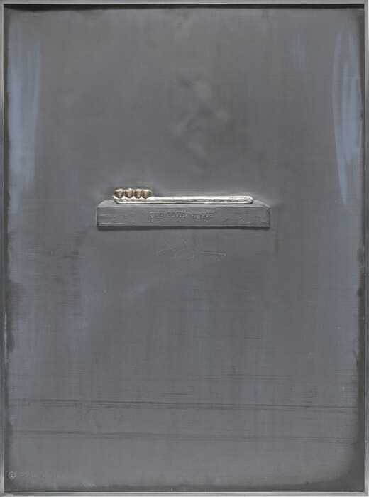A block holding a toothbrush set with gold teeth raised in low relief on a lead sheet. The rectangular block holding the brush is almost halfway down the panel. The brush has a straight, toothbrush-like handle highlighted with shiny silver tin leaf. Four gold teeth are set in a row at what would be the brush end, to our left. Letters are stamped in the front face of the block to read, “THE CRITIC SMILES.” The artist signed and dated the work below the block with dotted lines, “J Johns ’69.” Also incised in the lower left corner of the lead sheet is “GEMINI III” with a Gemini horoscope symbol.