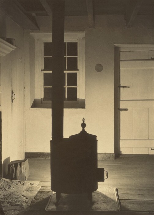A freestanding stove and pipe leading to the ceiling are backlit against the light-colored walls of a cabin in this vertical, black and white photograph. The panes of a window on the far wall are darkened, and both the upper and lower halves of the Dutch door are closed to the right. Light, presumably from the stove opening on the opposite side, rakes across the floor but leaves the beamed and timber ceiling veiled in shadow. A stone hearth to our left suggests a fireplace is just out of view to our left. The stove stands on a square tray and is topped with a teardrop-shaped finial.