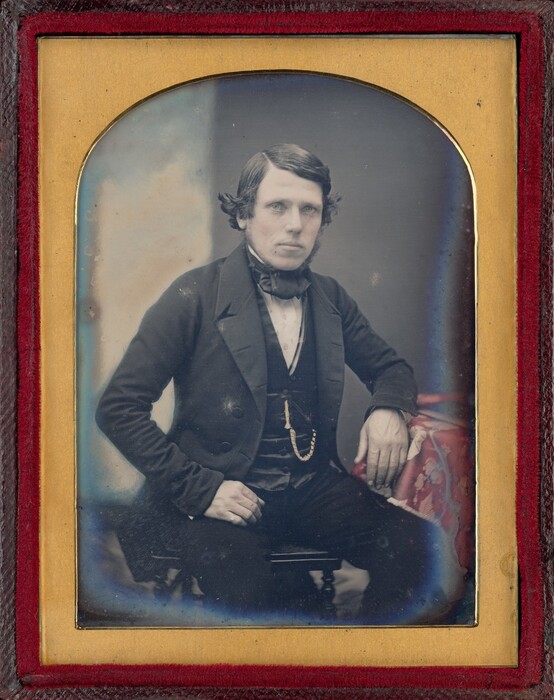 The image shows a man seated from the waist up. He has short, dark hair and styled sideburns. He is wearing a dark suit jacket, waistcoat, and a cravat. His left arm is resting on a table with a patterned cloth, and his right hand is on his thigh. The man's gaze is directed towards the camera, and he has a neutral expression. A watch chain hangs from his waistcoat. The image is framed in gold within a red frame, with a plain, neutral background typical of vintage photographs.