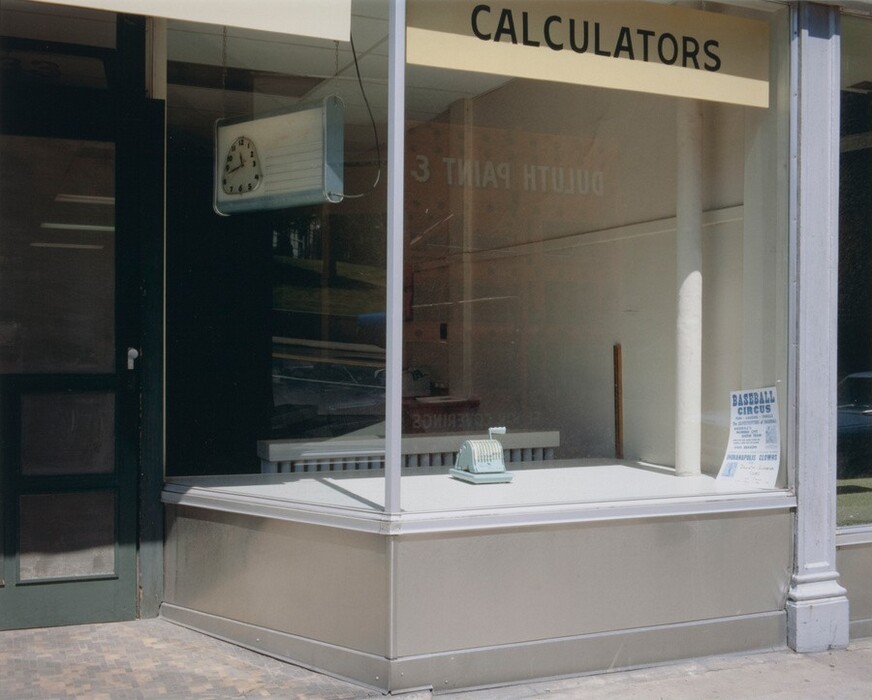 The image displays a storefront window with a small mint green vintage cash register and a sign advertising a "Baseball Circus" event. Soft pastel colors dominate the color palette, including the mint green of the cash register and the light beige or cream of the window frame and sign. The cash register is centered on a raised display platform in the middle of the window, while the sign is affixed to the lower right corner of the glass pane. Above the window, there is a beige-yellow banner with the word "CALCULATORS" in black letters. A retro-style clock is hanging inside on the left side, visible through the glass.