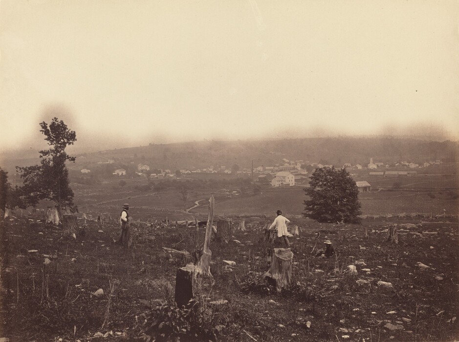 This sepia-toned photograph depicts a rural landscape littered with tree stumps and debris. Two people stand among the stumps, their backs to us. The person on the right wears a long light coat and a dark hat, and the person on the left wears a white shirt with a dark waistcoat and a pale gray hat and trousers. Two trees with dark leaves are visible, one on the left side and one to the right of center. In the distance is a low hill covered with white buildings. A dirt pathway winds through the landscape in the distance, and wooden fences cut through wide fields. The horizon is visible above the hills, which appear slightly obscured by light mist or fog. The sky is off-white.