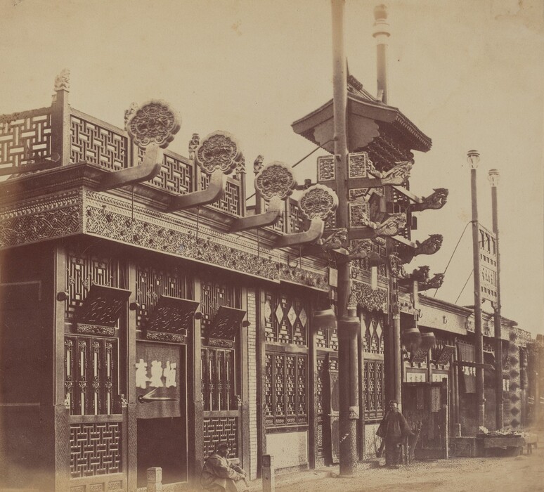 The image depicts a street scene with an ornate architectural facade. The perspective is from street level, looking directly at the building. The building has detailed latticework, carvings, and dragon-like sculptures. The color palette is sepia. In the foreground, two people are seated, partially in shadows. The street continues with similar architectural features in the distance. The image captures fine details typical of early photography.