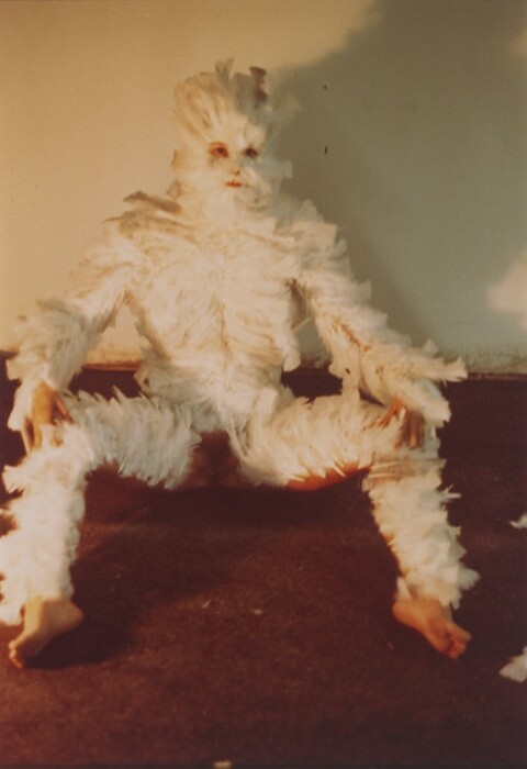 A person almost entirely covered with white feathers sits on a dark brown floor against a white wall in this vertical color photograph. The feathers leave the eyes, fingers, bare feet, and genitals visible. Woman sits with hands on knees, which are spread wide. She is lit from the left and casts a dark shadow on the wall to our right. The photograph is slightly out of focus.